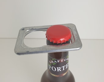 May include: A silver bottle opener with a red bottle cap on top. The bottle opener is shaped like a rectangle with a round hole in the center. The bottle cap is on top of a brown glass bottle with a label that says "Kraftwerk Porter".