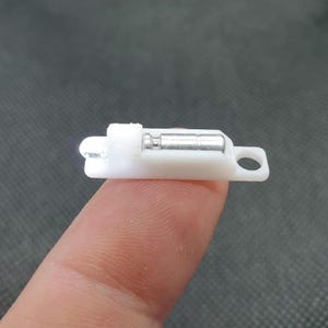 Może przedstawiać: Mała, biała plastikowa zatrzask z srebrnym metalowym cylindrem i małym otworem na jednym końcu. Zatrzask jest przeznaczony do zabezpieczania małych drzwi lub panelu. Obiekt jest trzymany przez palec.