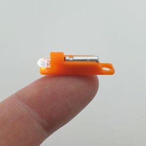 Könnte beinhalten: Eine orangefarbene LED-Leuchte mit einer klaren, abgerundeten Glühbirne und einer silberfarbenen Metallkomponente. Das Licht wird auf einem Finger gehalten, was seine geringe Größe und sein Design zeigt. Das Licht hat ein kleines Loch am Ende.