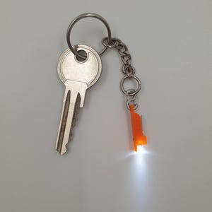 Może przedstawiać: Srebrny klucz przymocowany do breloczka z małą pomarańczową latarką LED. Latarka jest podświetlona, emitując jasne, białe światło. Breloczek zawiera mały łańcuszek i kółko na klucze.