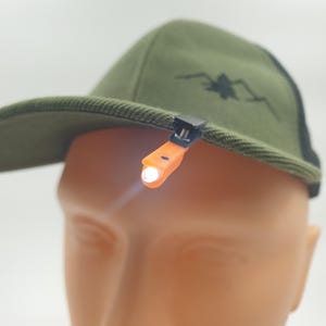 May include: An olive green baseball cap with a black mesh back and a small black graphic. An orange LED light is clipped to the brim, emitting a bright white beam. The light is attached to the cap with a black clip.