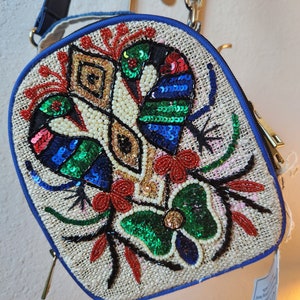 May include: A small, round, beige bag with a blue trim. The bag is decorated with colorful sequins and beads in the shape of flowers and birds. The bag has a gold zipper and a gold chain strap.