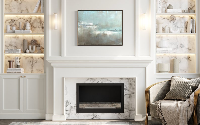 May include: A white fireplace with a black firebox and a marble surround. The fireplace is flanked by white built-in shelves with marble backs and gold accents. A light blue abstract painting hangs above the fireplace. A gold and white armchair with a gray throw and pillow sits in front of the fireplace.