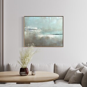 May include: A light blue and gray abstract painting in a gold frame hangs on a white wall above a light brown wooden table. A vase with white flowers sits on the table.