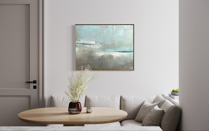 May include: A light blue and gray abstract painting in a gold frame hangs on a white wall above a light brown wooden table. A vase with white flowers sits on the table.