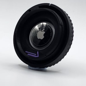 May include: Black camera lens with a silver Apple logo in the center. The lens has a purple inscription on the side.