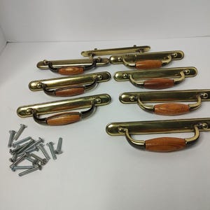 May include: A set of six brass drawer pulls with wooden center grips and matching metal mounting screws. Each pull features a long, rectangular brass backplate with rounded ends and a curved handle attached to the center, showcasing a wood grain finish.