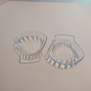 May include: Two clear glass seashell-shaped dishes. The dishes have a ridged design and are open at the top. One dish is upright, and the other is upside down. The dishes are on a white surface.