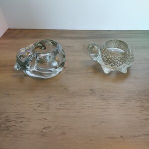 May include: Two clear glass candle holders. One is shaped like a cat, the other like a turtle. Both have a circular opening to hold a candle. The cat candle holder is lying on its side. The turtle candle holder is upright.