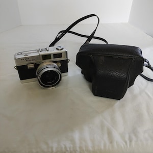 May include: A vintage Petri Racer camera with a black and silver body, lens, and strap. A black leather camera case sits next to the camera. The camera is on a white surface.