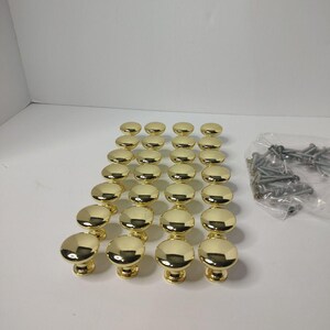 May include: A set of sixteen polished gold-tone metal cabinet knobs arranged in a four-by-four grid. Each knob features a smooth, rounded, mushroom-shaped head with a short cylindrical base. A clear plastic bag containing metal screws is included.