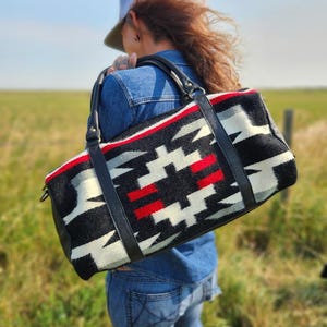 Puede incluir: Una bolsa de viaje de lona negra y blanca con detalles rojos. La bolsa tiene asas de cuero negro y la lleva una persona que lleva una chaqueta vaquera y pantalones vaqueros.