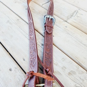 Puede incluir: Una cabezada de caballo de cuero marrón con una hebilla plateada y tallado floral. La cabezada está atada con una correa de cuero marrón.