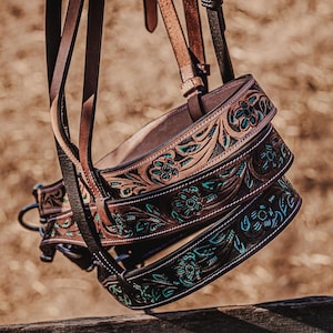 Puede incluir: Primer plano de una brida de caballo de cuero marrón con intrincados grabados florales y detalles turquesas. La brida cuelga de una valla de madera, mostrando la artesanía detallada. El cuero es de color marrón intenso.