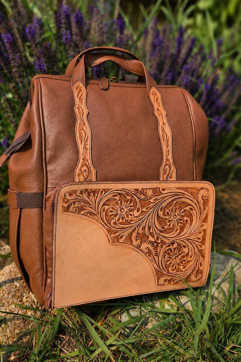K&ouml;nnte beinhalten: Ein brauner Lederrucksack mit einem Tragegriff und einer Vordertasche mit einer hellbraunen Lederblende. Die Frontblende und die vertikalen Riemen sind mit detaillierten Blumenpr&auml;gungen verziert. Der Rucksack verf&uuml;gt &uuml;ber einen Rei&szlig;verschluss.