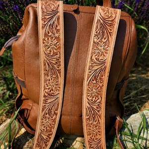 K&ouml;nnte beinhalten: Ein brauner Lederrucksack mit zwei breiten Schultergurten, die mit aufwendigen Blumenmustern verziert sind. Die hellbraunen Riemen zeigen gepr&auml;gte Bl&uuml;ten- und Blattmotive, die einen Kontrast zum glatten, mittelbraunen Leder des Rucksackk&ouml;rpers bilden.