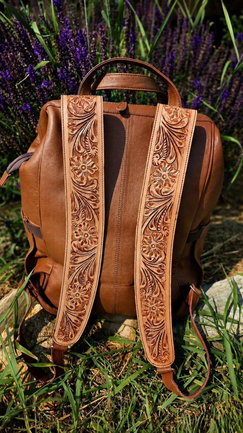 K&ouml;nnte beinhalten: Ein brauner Lederrucksack mit zwei breiten Schultergurten, die mit aufwendigen Blumenmustern verziert sind. Die hellbraunen Riemen zeigen gepr&auml;gte Bl&uuml;ten- und Blattmotive, die einen Kontrast zum glatten, mittelbraunen Leder des Rucksackk&ouml;rpers bilden.