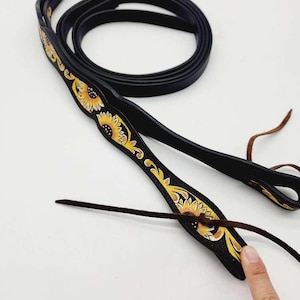 Può includere: Redini per cavalli in pelle nera con un motivo a girasole giallo e arancione. Le redini hanno un design ad anello e un laccio in pelle marrone. Il motivo a girasole è sulla parte superiore delle redini.