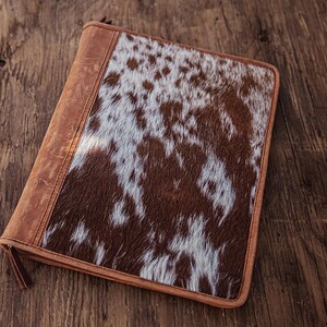 Puede incluir: Un portafolio rectangular de cuero con un patrón de piel de vaca. La cubierta presenta un diseño manchado marrón y blanco, con una lomo y ribete de cuero marrón. El portafolio se cierra con una cremallera y tiene un tirador de cuero.