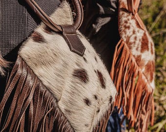 Könnte beinhalten: Nahaufnahme von zwei Kuhfell-Handtaschen. Die erste Tasche hat ein geflecktes Muster mit braunen Fransen und einem braunen Griff. Die zweite Tasche hat ein ähnliches Design mit einem anderen gefleckten Muster und braunen Fransen.