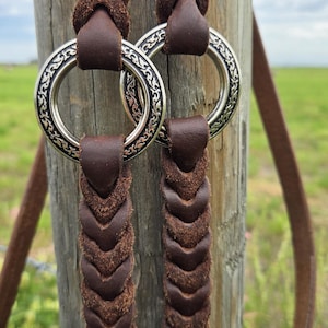 May include: Two dark brown braided leather straps connected by two silver-toned metal rings. Each ring features an intricate embossed pattern along its outer edge. The leather has a textured, matte finish and a consistent braided weave pattern.