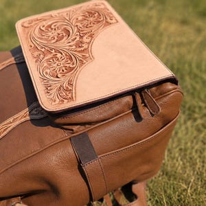 K&ouml;nnte beinhalten: Ein brauner Lederrucksack mit einer hellbeigen Lederklappe, die mit aufwendigen floralen Pr&auml;gungen versehen ist. Der Rucksack hat eine genarbte Oberfl&auml;che, passende Lederriemen mit Verzierungen und einen Rei&szlig;verschluss mit zwei Zippern.