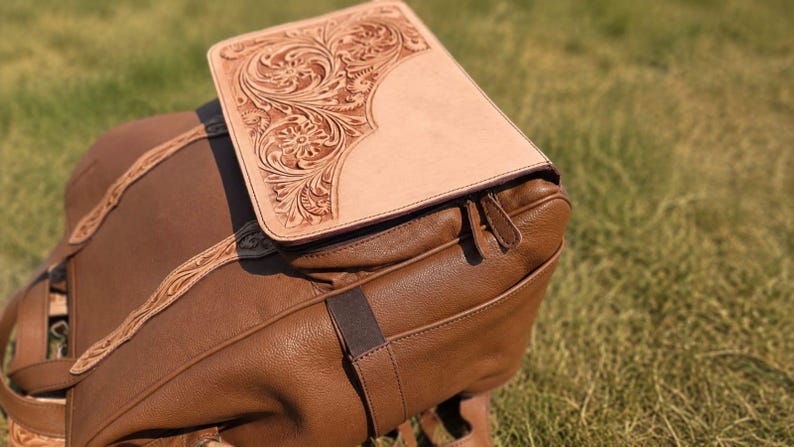 K&ouml;nnte beinhalten: Ein brauner Lederrucksack mit einer hellbeigen Lederklappe, die mit aufwendigen floralen Pr&auml;gungen versehen ist. Der Rucksack hat eine genarbte Oberfl&auml;che, passende Lederriemen mit Verzierungen und einen Rei&szlig;verschluss mit zwei Zippern.