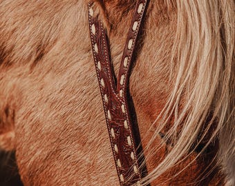 May include: A dark brown leather horse headstall featuring intricate floral tooling and light-colored rawhide lacing along the edges. The headstall is positioned on the side of a light brown horse with a blonde mane, showcasing the detailed craftsmanship.