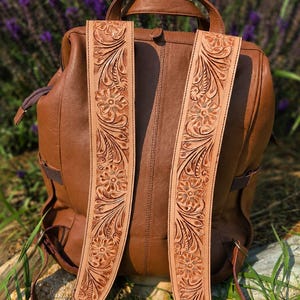 K&ouml;nnte beinhalten: Ein brauner Lederrucksack mit zwei breiten Schultergurten, die mit detaillierten, hellen Blumen- und Blattpr&auml;gungen verziert sind. Der Rucksack verf&uuml;gt &uuml;ber einen Tragegriff, ein Hauptfach mit Rei&szlig;verschluss sowie seitliche Rei&szlig;verschl&uuml;sse.