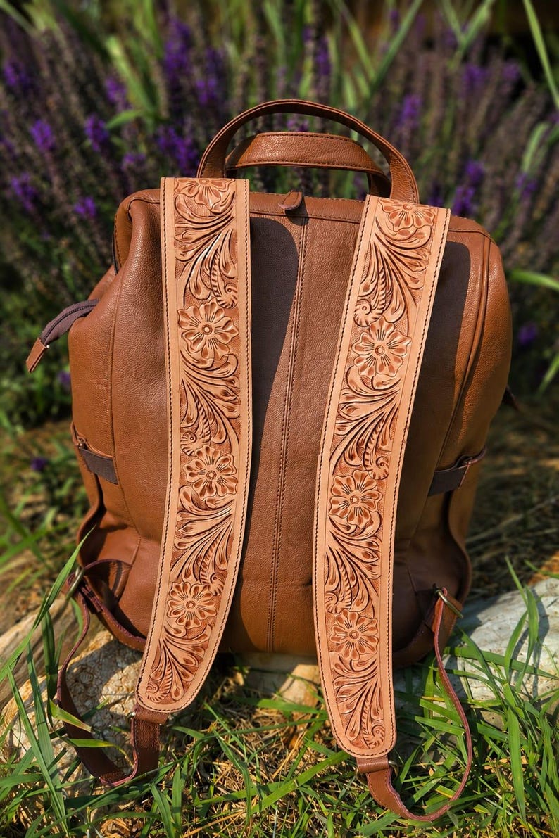 K&ouml;nnte beinhalten: Ein brauner Lederrucksack mit zwei breiten Schultergurten, die mit detaillierten, hellen Blumen- und Blattpr&auml;gungen verziert sind. Der Rucksack verf&uuml;gt &uuml;ber einen Tragegriff, ein Hauptfach mit Rei&szlig;verschluss sowie seitliche Rei&szlig;verschl&uuml;sse.