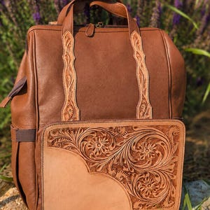 K&ouml;nnte beinhalten: Ein brauner Lederrucksack mit einem Tragegriff und zwei vertikalen Riemen mit kunstvoller Blumenpr&auml;gung. Die Vordertasche ist mit einem gro&szlig;en, rechteckigen Einsatz aus hellbraunem Leder verziert, das detaillierte Blumenmuster zeigt.