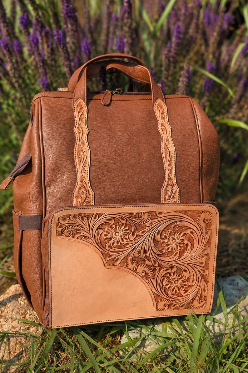 K&ouml;nnte beinhalten: Ein brauner Lederrucksack mit einem Tragegriff und zwei vertikalen Riemen mit kunstvoller Blumenpr&auml;gung. Die Vordertasche ist mit einem gro&szlig;en, rechteckigen Einsatz aus hellbraunem Leder verziert, das detaillierte Blumenmuster zeigt.