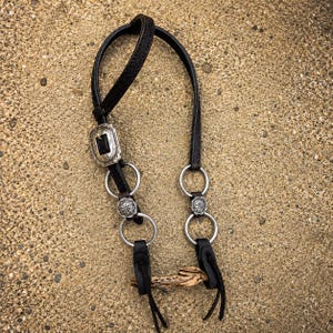 May include: A dark brown leather horse bridle component featuring silver-toned metal hardware, including a rectangular buckle with a black inlay, circular rings, and decorative floral-patterned conchos, ending in two leather tie-down straps.