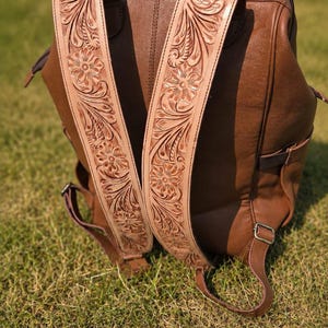 K&ouml;nnte beinhalten: Ein brauner Lederrucksack mit breiten, hellen Schultergurten, die mit detaillierten Blumenpr&auml;gungen und Rankenmustern verziert sind. Der Hauptteil des Rucksacks besteht aus glattem, braunem Leder mit einem Tragegriff und Metallbeschl&auml;gen.