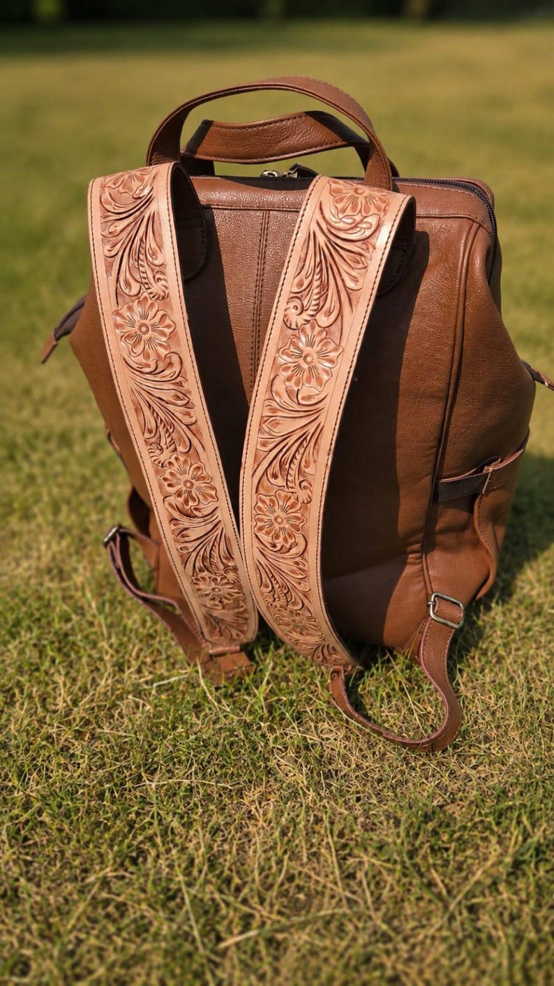 K&ouml;nnte beinhalten: Ein brauner Lederrucksack mit breiten, hellen Schultergurten, die mit detaillierten Blumenpr&auml;gungen und Rankenmustern verziert sind. Der Hauptteil des Rucksacks besteht aus glattem, braunem Leder mit einem Tragegriff und Metallbeschl&auml;gen.