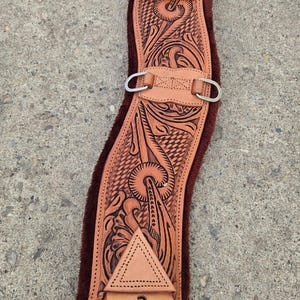 Può includere: Un sottopancia per cavallo in pelle marrone chiaro con intricati motivi floreali e geometrici intagliati. Il sottopancia ha una fodera in peluche marrone scuro e fibbie e ferramenta in metallo color argento. Il design è simmetrico e dettagliato.