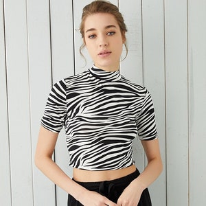 Puede incluir: Top corto con estampado de cebra en blanco y negro, con mangas cortas y cuello alto. Se combina con pantalones negros. El conjunto es informal y elegante.