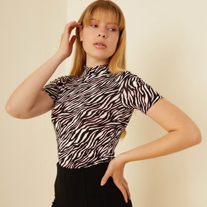Puede incluir: Una camiseta de cuello alto con estampado de cebra en blanco y negro con mangas cortas. La camiseta es ajustada y tiene un tejido elástico.
