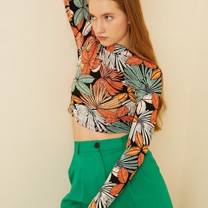 May include: A black long-sleeved shirt with a tropical leaf print in shades of orange, green, and white. The shirt is cropped and has a fitted silhouette. The model is wearing the shirt with green high-waisted pants.