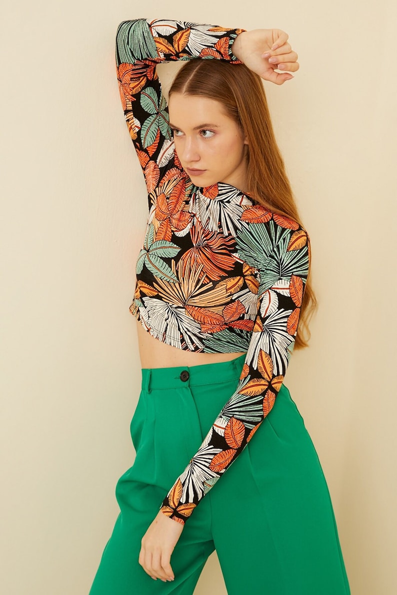May include: A black long-sleeved shirt with a tropical leaf print in shades of orange, green, and white. The shirt is cropped and has a fitted silhouette. The model is wearing the shirt with green high-waisted pants.