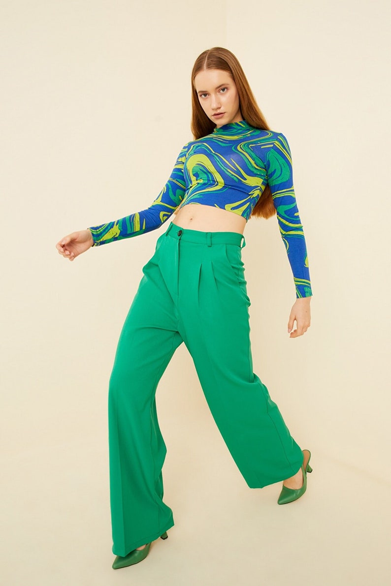 Pu&ograve; includere: Una donna indossa un top corto a maniche lunghe con stampa astratta blu e verde e pantaloni larghi verdi.