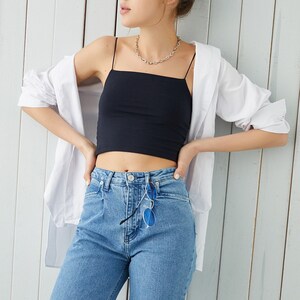 May include: A woman wearing a black crop top with thin straps, a white button-down shirt, and blue jeans. She is wearing a silver chain necklace and has her hair pulled back in a bun.