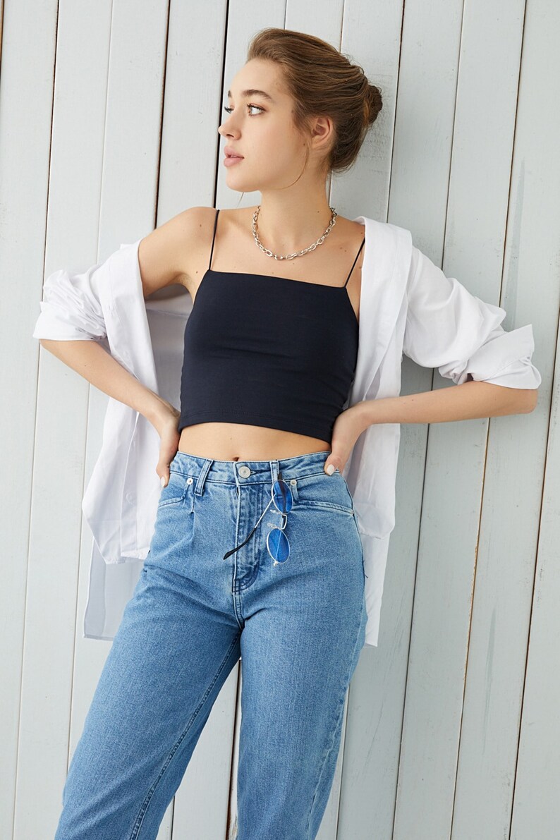 May include: A woman wearing a black crop top with thin straps, a white button-down shirt, and blue jeans. She is wearing a silver chain necklace and has her hair pulled back in a bun.