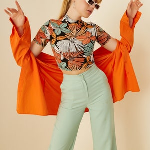 May include: A woman wearing a black and orange floral print crop top, a bright orange shirt, and light green wide-leg pants. She is wearing white rectangular sunglasses.