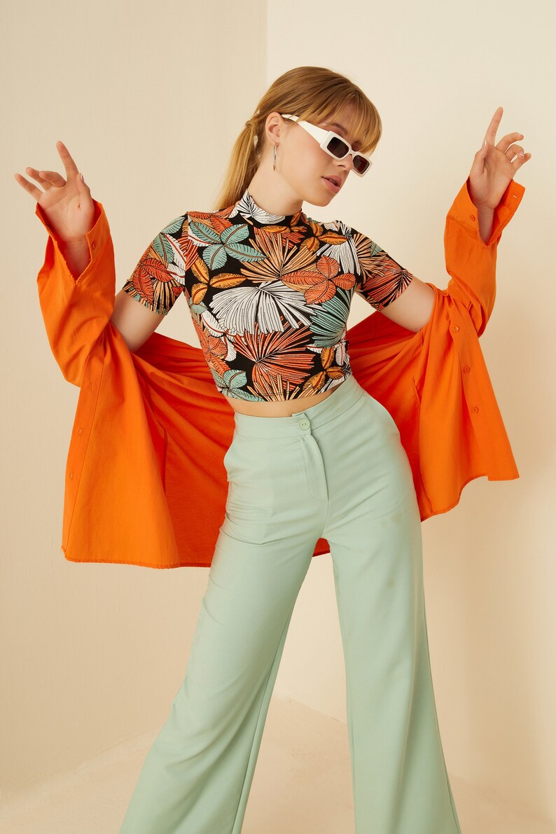 May include: A woman wearing a black and orange floral print crop top, a bright orange shirt, and light green wide-leg pants. She is wearing white rectangular sunglasses.