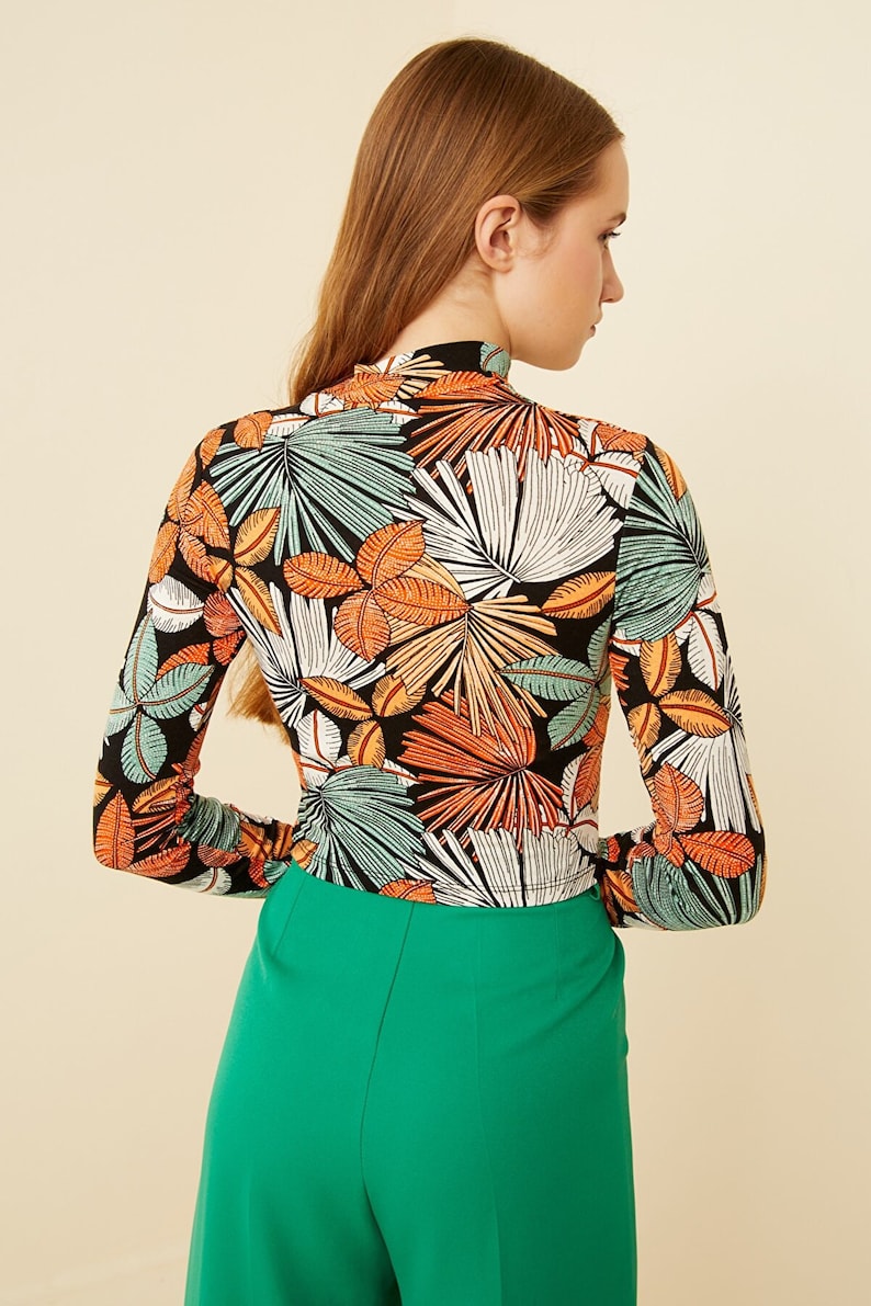 May include: A black long-sleeved shirt with a tropical leaf print in shades of green, orange, and white. The shirt has a mock neck and a fitted silhouette.