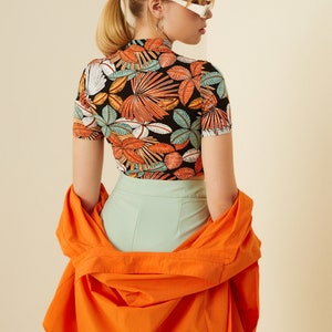 May include: A woman wearing a black and orange tropical print crop top with a high neck and short sleeves. She is also wearing a light green pair of pants and an orange shirt draped over her shoulders.