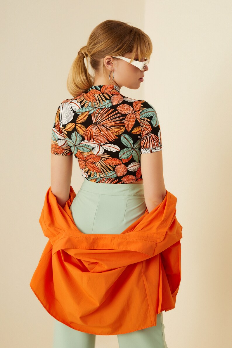 May include: A woman wearing a black and orange tropical print crop top with a high neck and short sleeves. She is also wearing a light green pair of pants and an orange shirt draped over her shoulders.