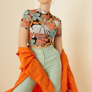 May include: A woman wearing a short-sleeved, black crop top with a colorful tropical print, light green wide-leg pants, and a bright orange button-down shirt. She is wearing white rectangular sunglasses.