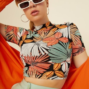 May include: A black crop top with a tropical leaf print. The top has a mock neck and short sleeves. The model is wearing the top with a pair of light green pants and a bright orange shirt.